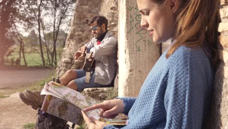 happy young couple backpackers tourists sitting near wooden doorway of medieval building in parco degli acquedotti park ruins in rome at sunrise with guitar and map guide slow motion steadycam