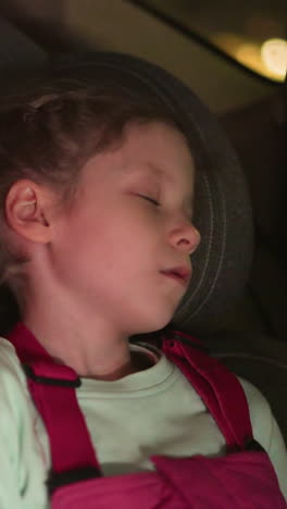 una vista de cerca de un niño durmiendo pacíficamente en un asiento de coche, iluminado por el brillo suave de las luces ambientales, el niño lleva un mono rosa y una camisa verde claro, descansando cómodamente