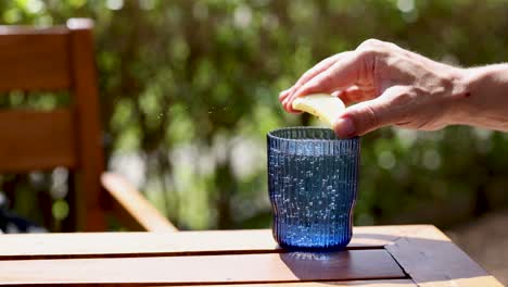 una mano exprime limón en un vaso azul en una mesa iluminada por el sol, creando una escena refrescante