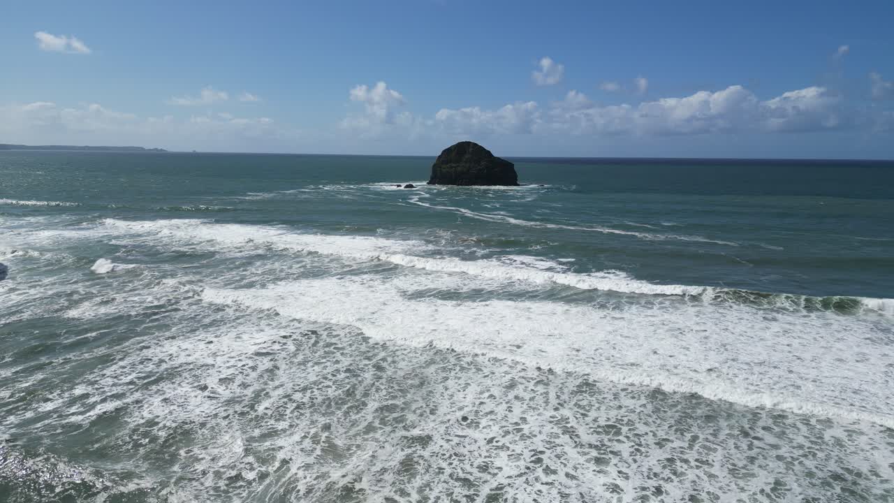 Aerial drone view of Trebarwith Strand beach in Cornwall, showing waves crashing on the shore with a large rock in the distance. The calm, blue waters and clear skies create a serene coastal scene