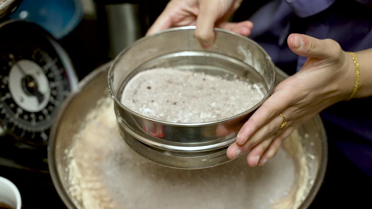 Hands skillfully shaking a sieve to mix flour and cocoa powder, creating a fine blend for baking, with a scale visible in the background