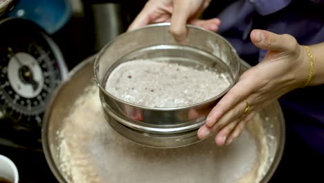 hands skillfully shaking a sieve to mix flour and cocoa powder, creating a fine blend for baking, with a scale visible in the background