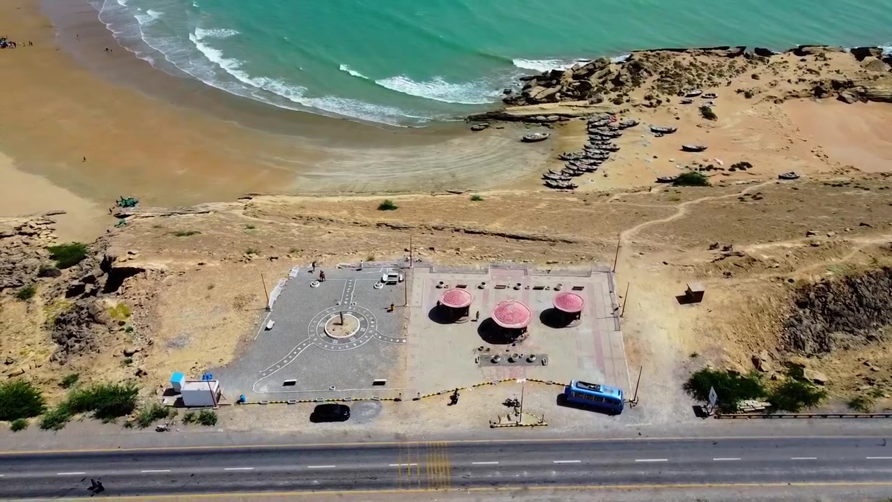 Video de dron captura una vista aérea panorámica del área de descanso junto a la carretera de Kund Malir y la playa turquesa en Baluchistán, mostrando colinas áridas iluminadas por el sol, vehículos estacionados y un paisaje costero tranquilo.