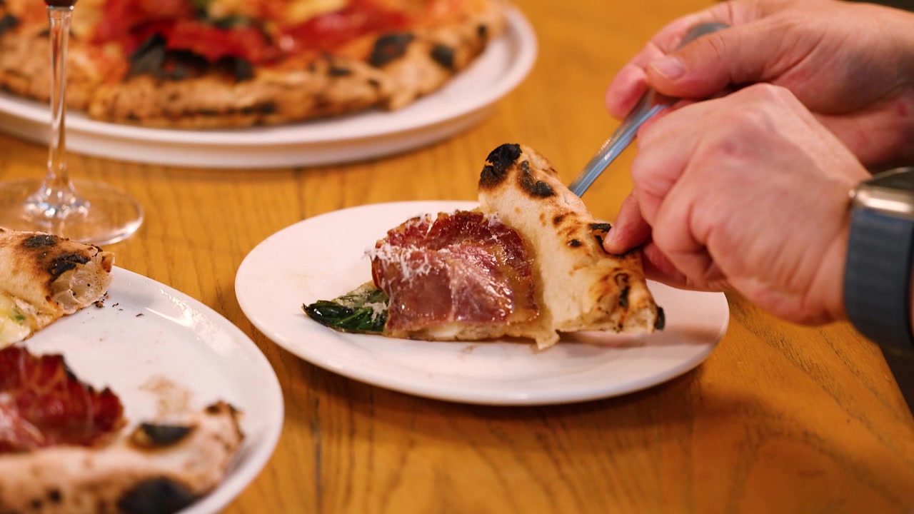 Primo piano di mani che usano una forchetta per spostare una fetta di pizza al prosciutto su un piatto.