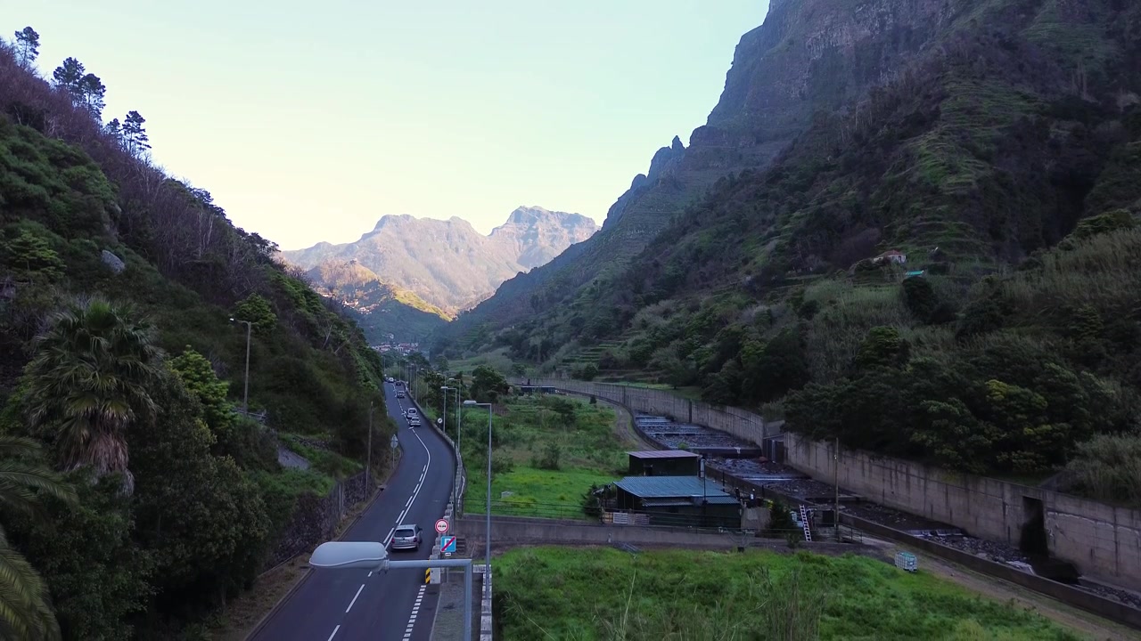 Riprese aeree con drone catturano una profonda gola di montagna sull'isola di Madeira con ripide pareti rocciose coperte da una vegetazione lussureggiante. Una strada tortuosa attraversa il fondovalle, evidenziando il terreno accidentato.