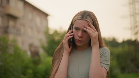 young woman showing exhaustion and frustration during a phone call, holding her left hand to her forehead, set against a blurred outdoor background of greenery and distant building