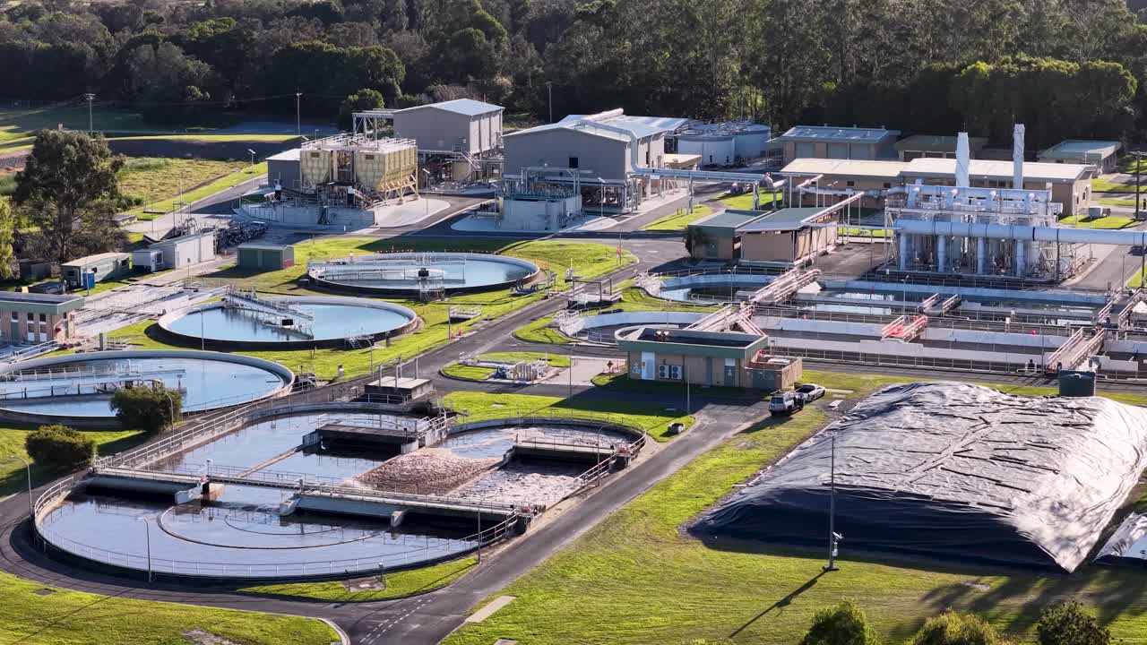 Aerial camera smoothly pans across a large wastewater treatment plant with circular tanks, green lawns, and industrial buildings under bright daylight