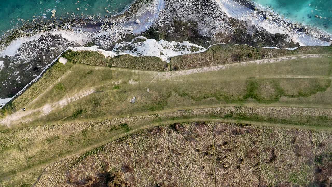Aerial cliff-top drop off reveals a bird’s-eye view of Beachy Head’s dramatic white cliffs, iconic red and white lighthouse, rocky beach, and calm waves for a stunning coastal perspective.