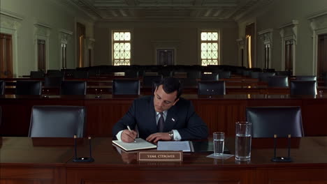 a man in a suit writing at a desk in a formal, empty meeting room
