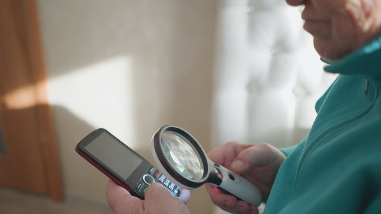 Elderly woman in green jacket holding mobile phone and magnifying glass, closely inspecting small details on screen, focused expression, bright indoor setting, sitting in comfortable chair, well-lit background