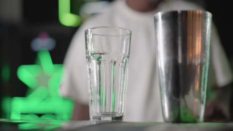 bartender wearing white shirt prepares drink, holding glass in hand in modern bar with green lighting in background. focused on drink preparation, bartending skills in vibrant environment