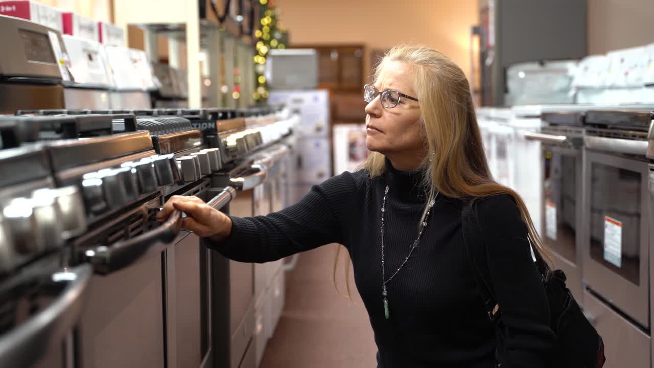 Belle femme blonde d’âge mûr regardant les fonctionnalités et les avantages d’une cuisinière à gaz dans un magasin d’électroménager