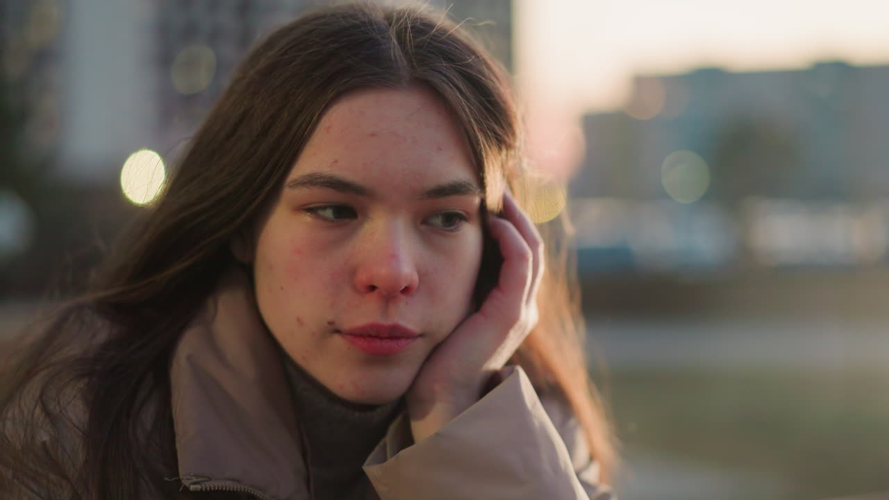 A close-up shot of a girl in a peach jacket, lost in deep thought as she gently rests her cheek on her hand. A faint smile graces her face. The blurred background with evening lights