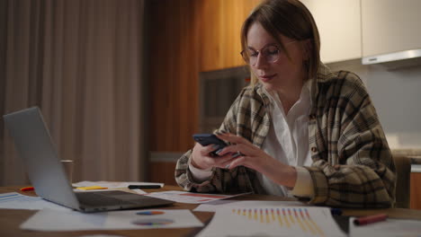 Glückliche-Frau,-Die-Ihr-Mobiltelefon-Benutzt,-Während-Sie-Zu-Hause-Mit-Einem-Laptop-Arbeitet.-Lächelnde,-Coole-Frau-Mit-Brille,-Die-Nachrichten-Mit-Dem-Smartphone-Sendet.-Schöne,-Stilvolle-Dame,-Die-Auf-Dem-Handy-Auf-Der-Website-Surft