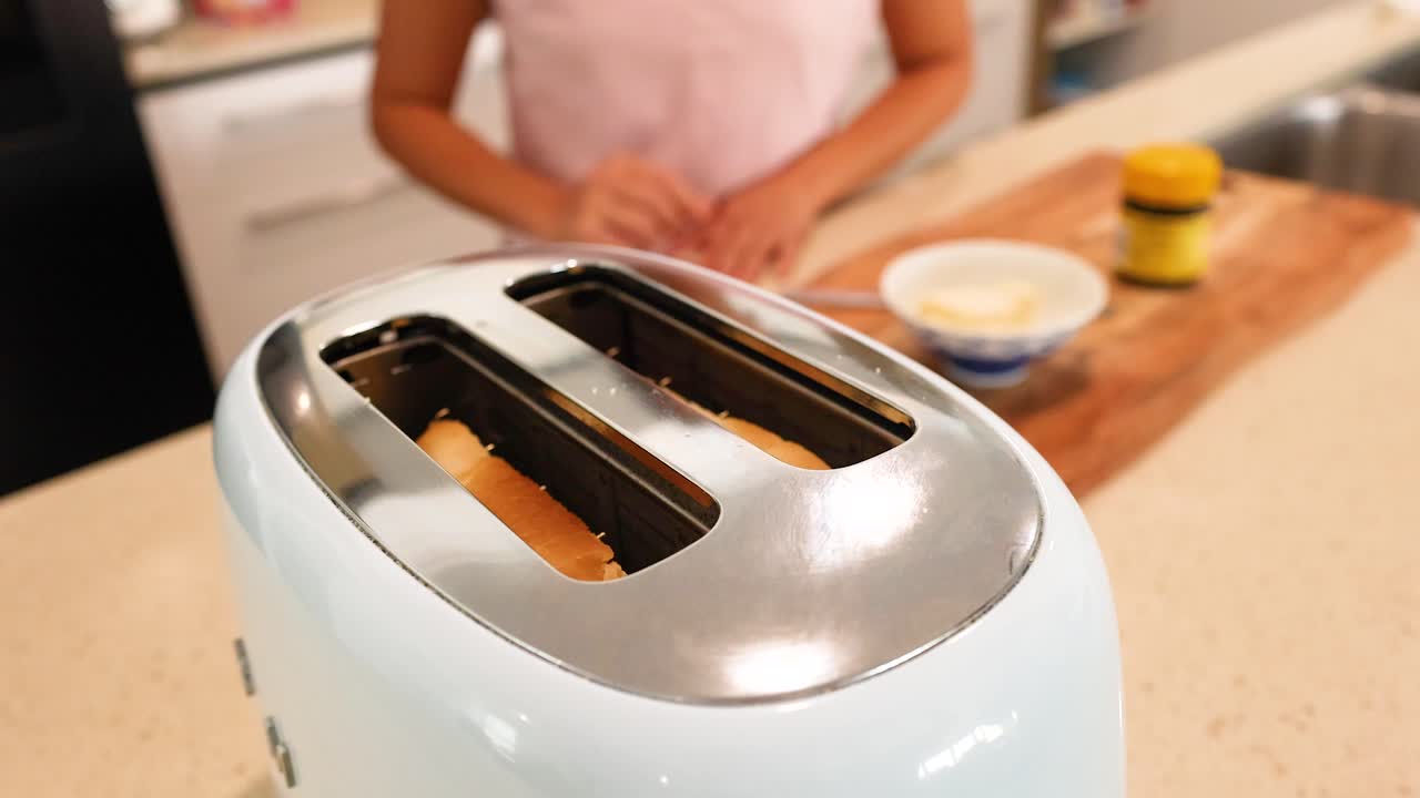 A person prepares toast in a bright kitchen, focusing on a toaster and bread. Warm lighting enhances the cozy atmosphere