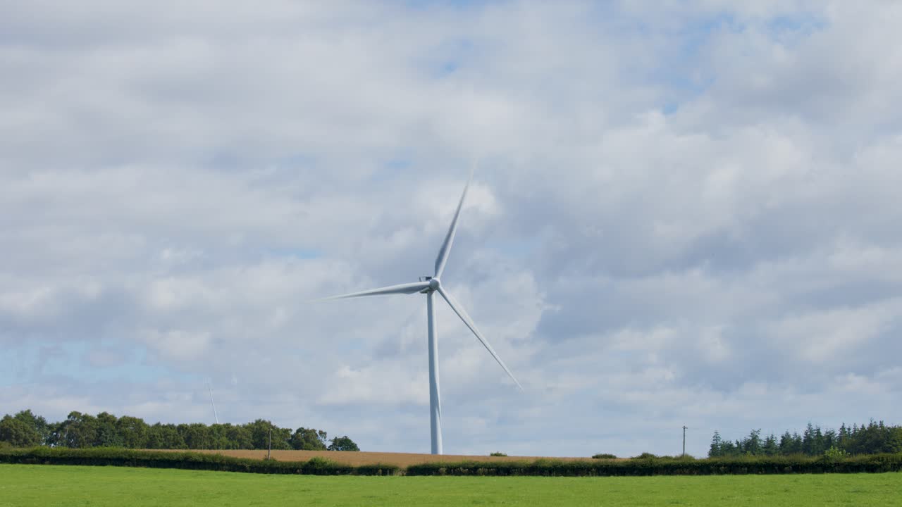 A single wind turbine spins steadily in a green rural landscape near Edinburgh, Scotland. Daylight, soft natural lighting, and a static wide shot capture the serene environment