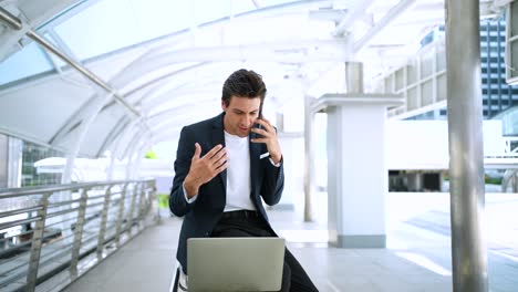 confident businessman using laptop working outdoors in the financial district in the city. using phone talking to client solving problems