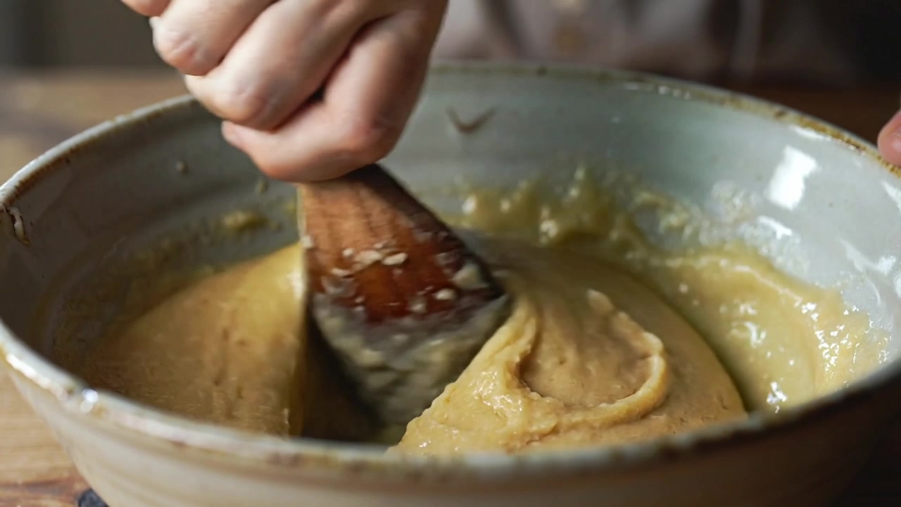 Pressing wooden spoon, cook's hands folding batter in ceramic bowl on wooden countertop for baking
