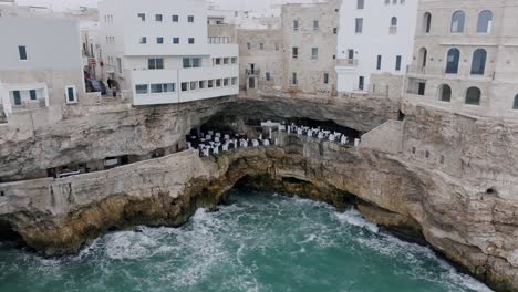 Imágenes-Aéreas-De-Un-Restaurante-En-Polignano-Una-Yegua-Que-Se-Encuentra-Dentro-De-Una-Cueva-En-Los-Acantilados-Con-Las-Olas-Del-Mar-Mediterráneo-Rompiendo-Debajo