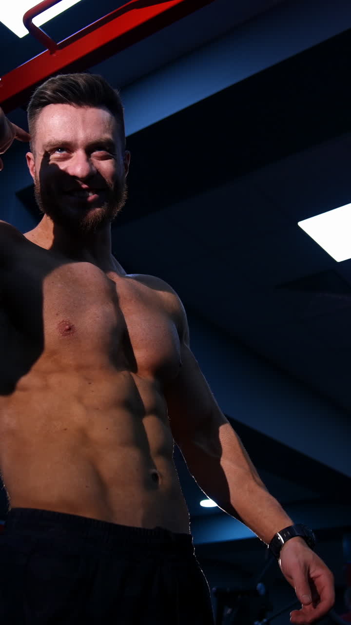 Healthy bodybuilder indoors. Smiling young man with sportive body talking in the modern gym. Handsome shirtless sportsman. View from below. Vertical video
