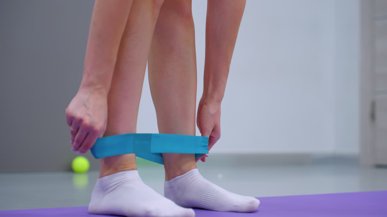 보라색 매트 위에 색 양말을 입고 발목 주위에 파란색 저항 밴드를 부착하는 사람의 클로즈업 테니스 공이 배경에 부드럽게 흐릿합니다.