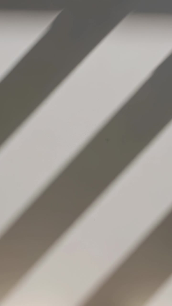 Vertical video: Casting light through blinds, diagonal dark-gray bands drifting across neutral wall