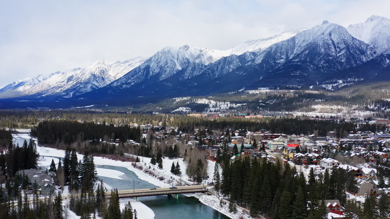 A drone flies into Canmore, revealing a vibrant town blanketed in snow and framed by the towering BANFF mountain range in Canada.