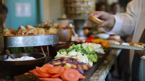 smooth shot of a line of people at a crowded event going through a food line and grabbing a variety of food including meats, cheeses, bread, fruits, and vegetables