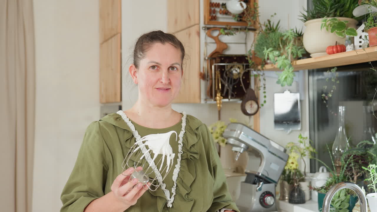 House wife in green apron stands in bright kitchen holding stainless mixer coated with thick whipped flour as she talks with cheerful expression surrounded by cozy rustic decor and plants
