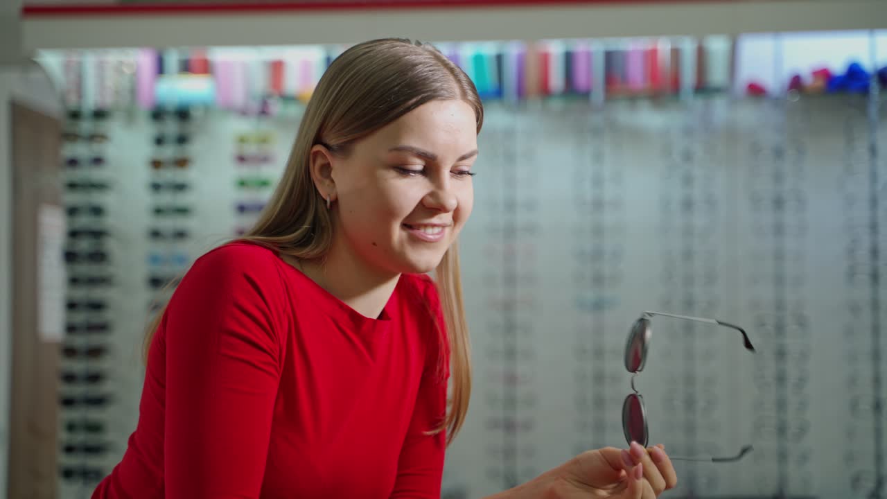Young woman trying on stylish sunglasses. Pretty girl in red blouse dresses black eyeglasses indoors. Eyesight concept.
