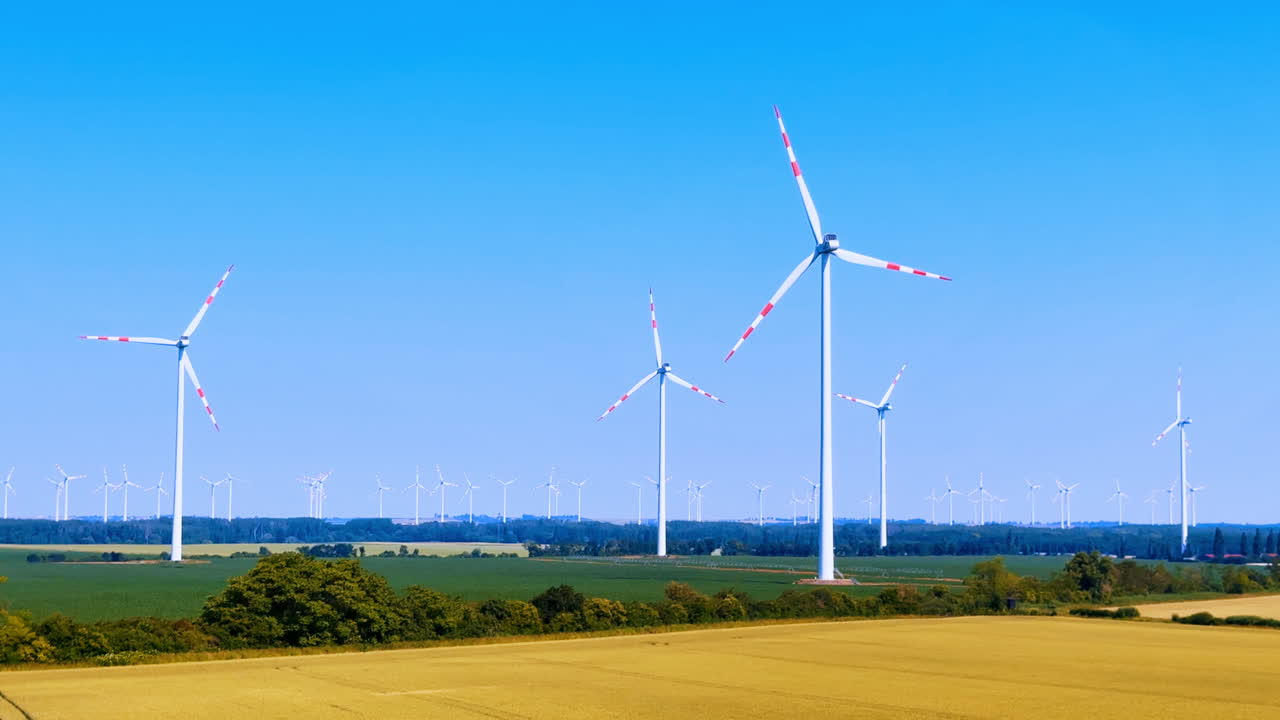 Turbine eoliche si ergono in un campo verde. Le turbine eoliche girano lentamente in un grande campo sotto un cielo sereno. La vista mostra terreni agricoli e alberi in lontananza.