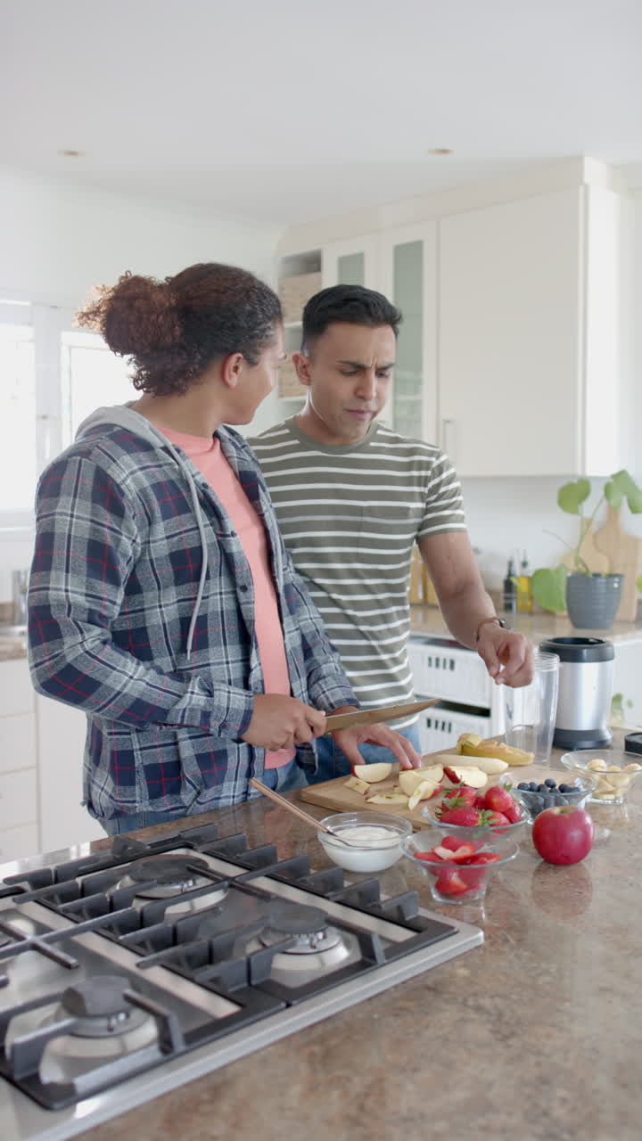 Vertical video of happy diverse gay male couple making fruit smoothie in kitchen, slow motion