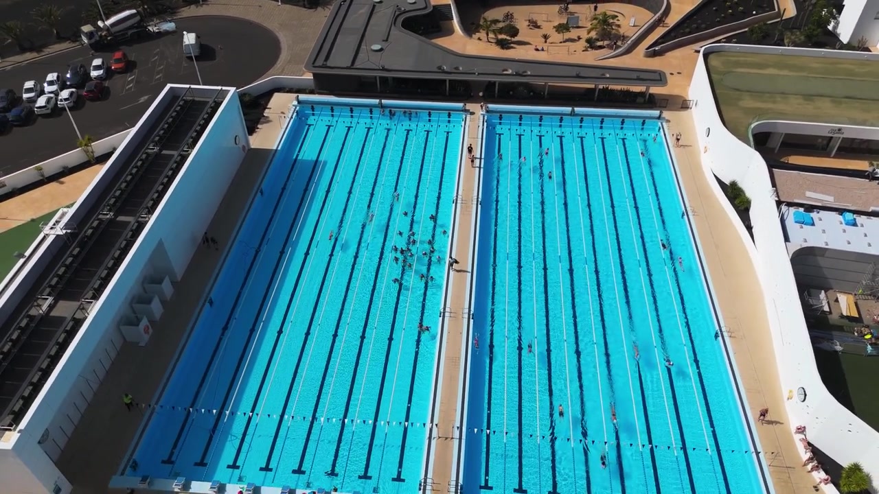 Two large outdoor lap pools with surfers and marked lanes in a sports complex, aerial establishing orbit with surfers in class training