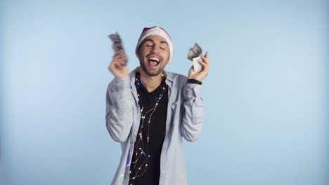 Excited-man-holding-paper-money,-dollar-banknotes.-Shaking-money-and-dancing-while-standing-against-the-blue-wall-in-santa-hat-and-garland-on-neck.-Get-money-cash,-receive-winner-payment,-gift-concept.-Man's-hand-hold-and-counting-cash-money-and-showing-in-excitement
