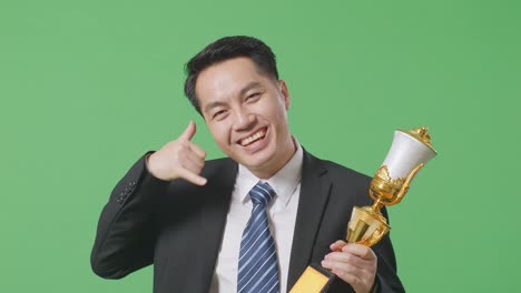 close up of asian business man in a suit and tie with a gold medal and trophy showing call me gesture and smiling to camera as the first winner on green screen background in the studio