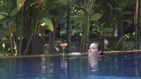 tiro largo de una mujer joven que se relaja al lado de la piscina mientras disfruta de un cóctel en un hotel tropical