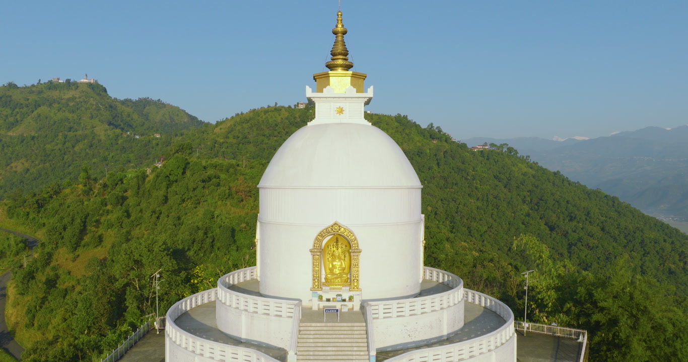 Aerial pull-in shot moving towards the white World Peace Stupa located high in the dense green forested mountains surrounding Pokhara Nepal.