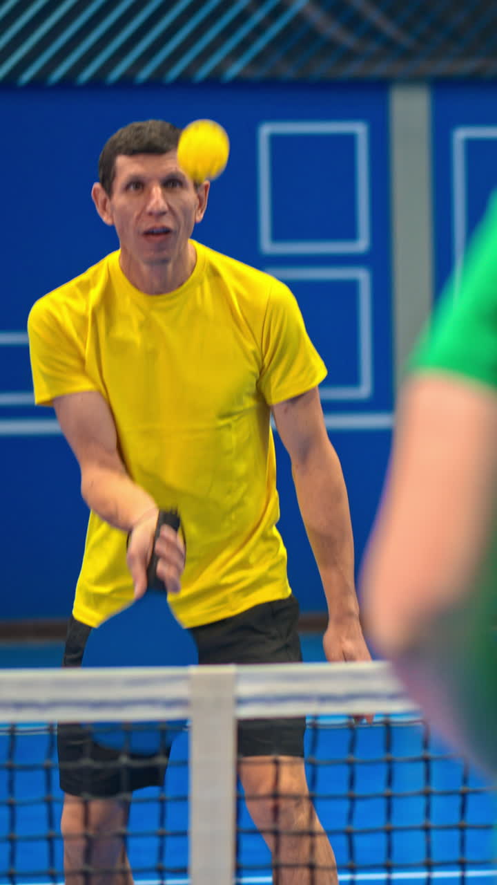 A man and a woman playing pickleball on a blue, inside court. Vertical
