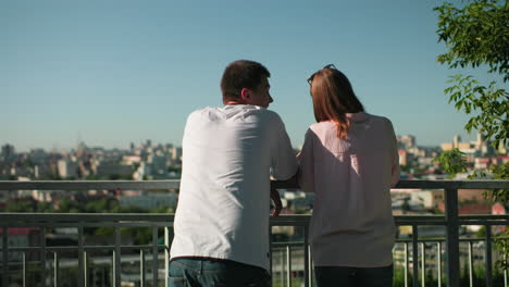 siblings leaning on iron railing interacting warmly with girl having her glasses on her head, background showcases a vibrant cityscape under clear skies