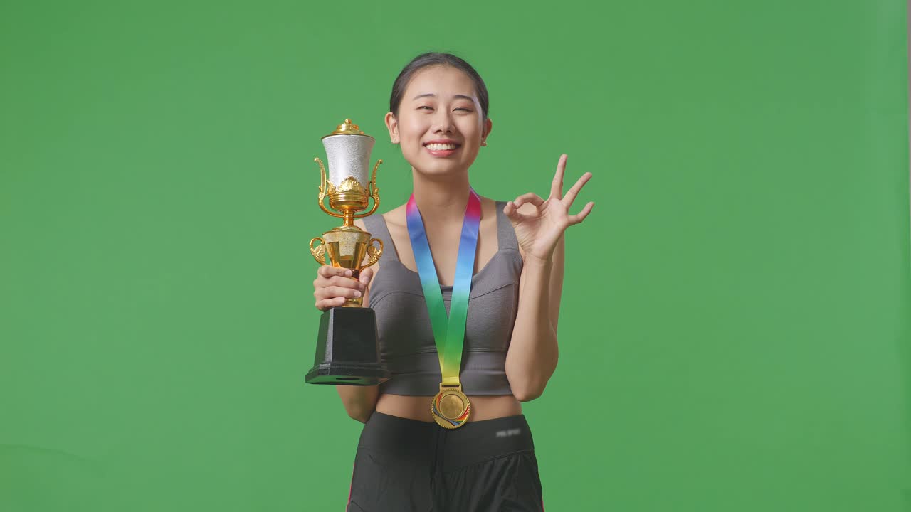 금메달과 트로피를 가진 아시아 여성은 스튜디오의 녹색 스크린 배경에서 첫 번째 우승자로 은 몸짓을 보여주고 카메라에 미소 짓고 있습니다.