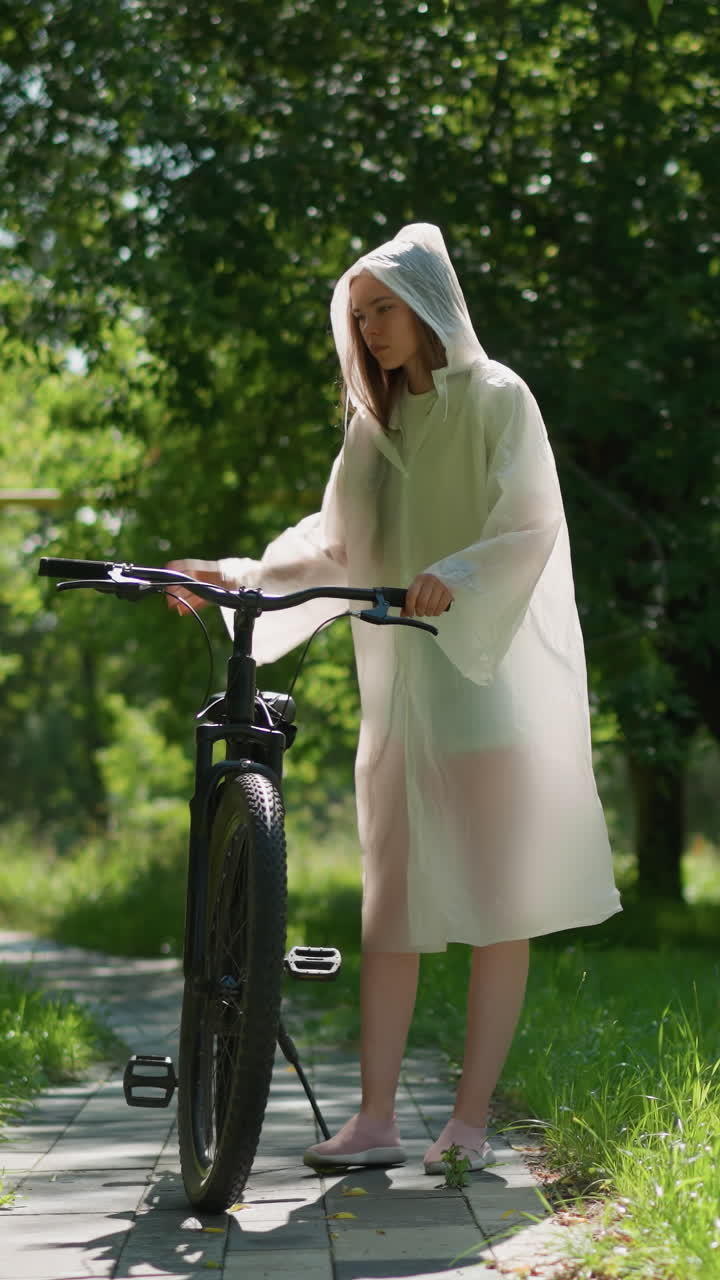 반투명한 레인코트를 입은 젊은 여자가 자전거를 타고 이 많은 길을 걸어가며 기둥 근처에 서서 멈추고, 후드를 내려놓고, 바닥에 기대어, 울창한 초록색과 나무로 둘러싸여 있습니다.