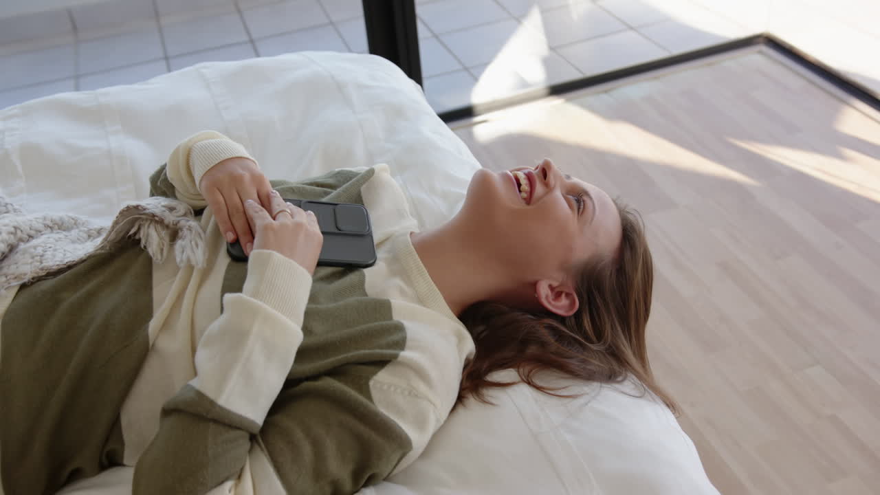 Relaxing on bed, young woman holding smartphone and smiling joyfully