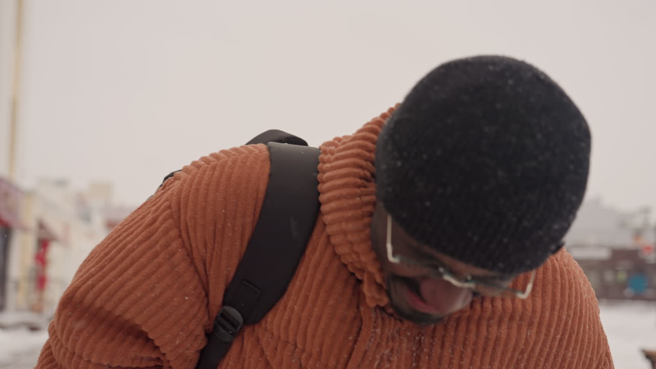 Black man smiling in snowy street wearing orange chunky sweater black beanie glasses and mittens with backpack gentle snowfall downtown shops blurred candid closeup capturing laughter cozy urban