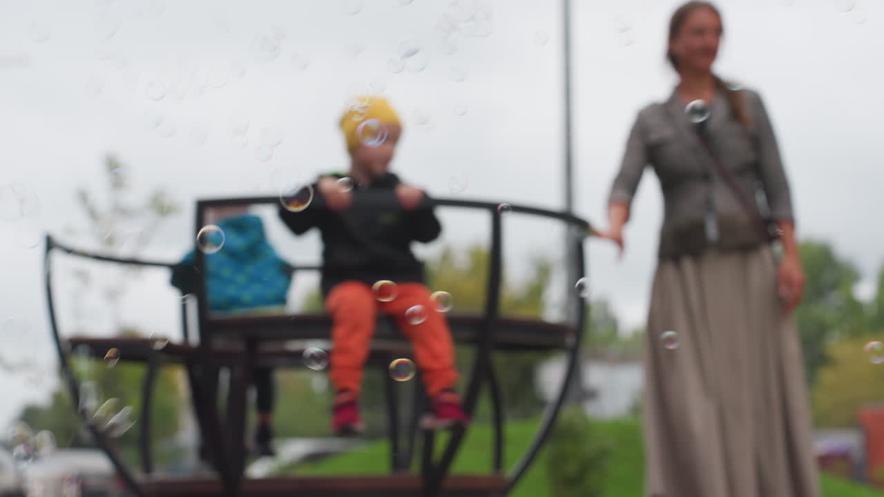 Blur background shows mother spinning two children on merry go round while soap bubbles float across frame, overcast sky above park, gentle motion and soft focus railings and seats