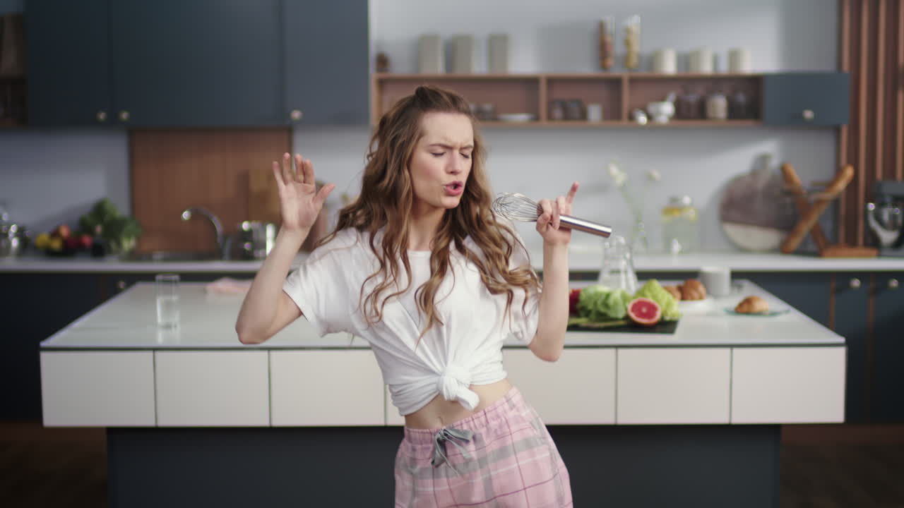 Donna sorridente che canta una canzone in una frusta in cucina. Ragazza allegra che si diverte.