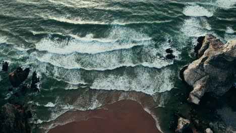 aerial view of a dramatic coastline with powerful waves crashing against rocks and a reddish-brown sandy beach.