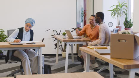 muslim businesswoman sitting at a table taking notes while an businesswoman talks to the new employee looking at laptop at another table in the office 1