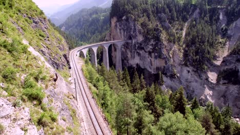 a stunning arched stone viaduct stretches across a deep gorge, surrounded by steep cliffs and lush green trees in the swiss alps on a clear day.