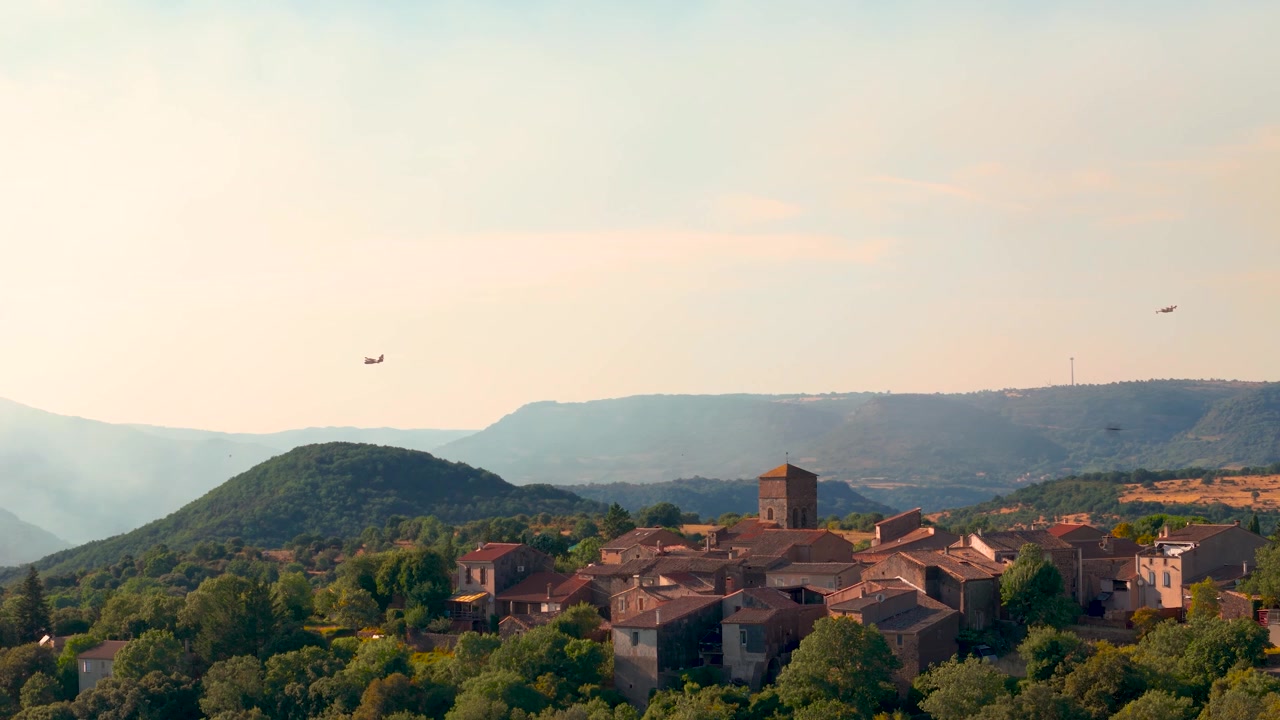 Vista aerea con drone di un villaggio nel sud della Francia in primo piano, con alberi e aerei Canadair che sorvolano l'area circostante per combattere vasti incendi boschivi, con fumo proveniente dagli incendi.