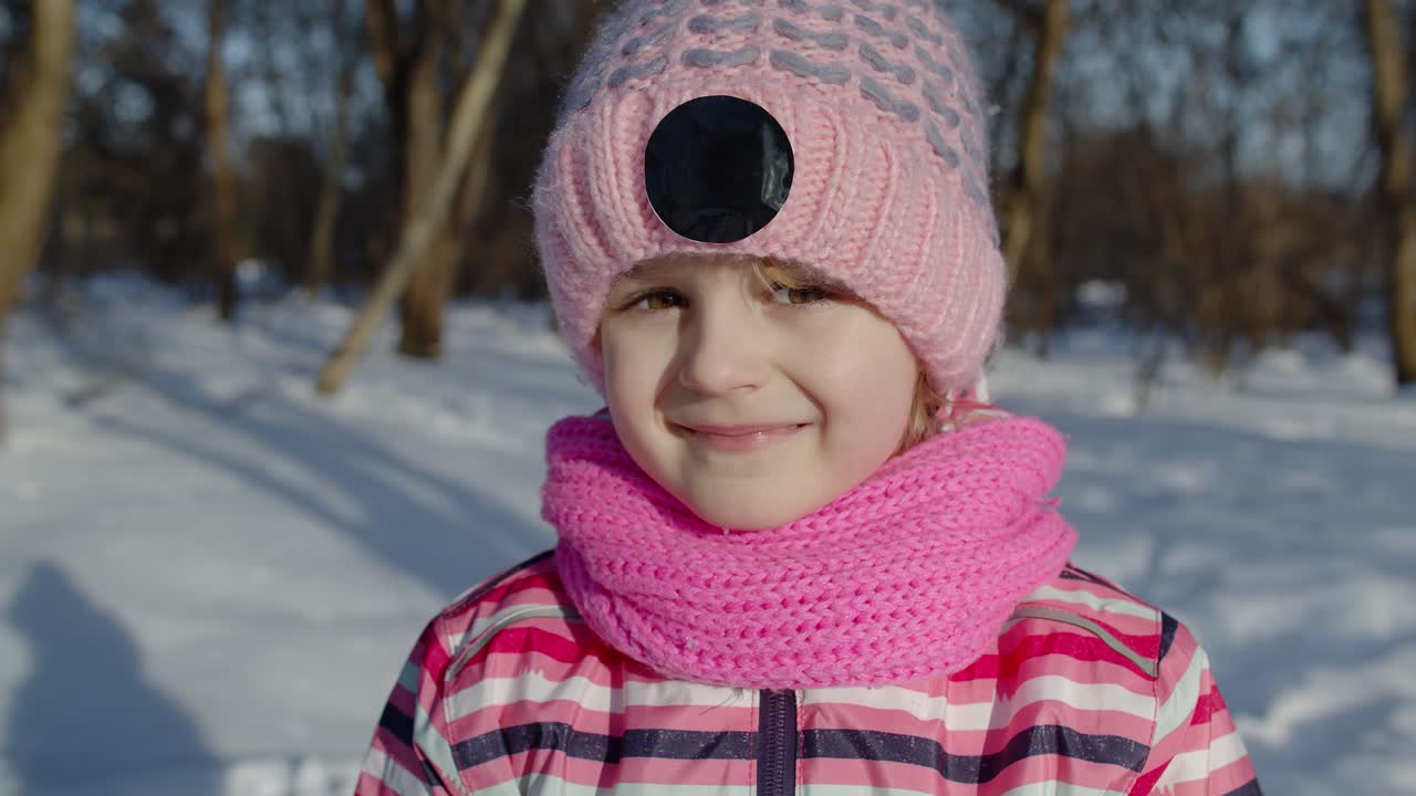 기쁜 어린 소녀가 카메라를 쳐다보고, 미소 짓고, 놀고, 겨울 공원에서 얼굴을 짓고